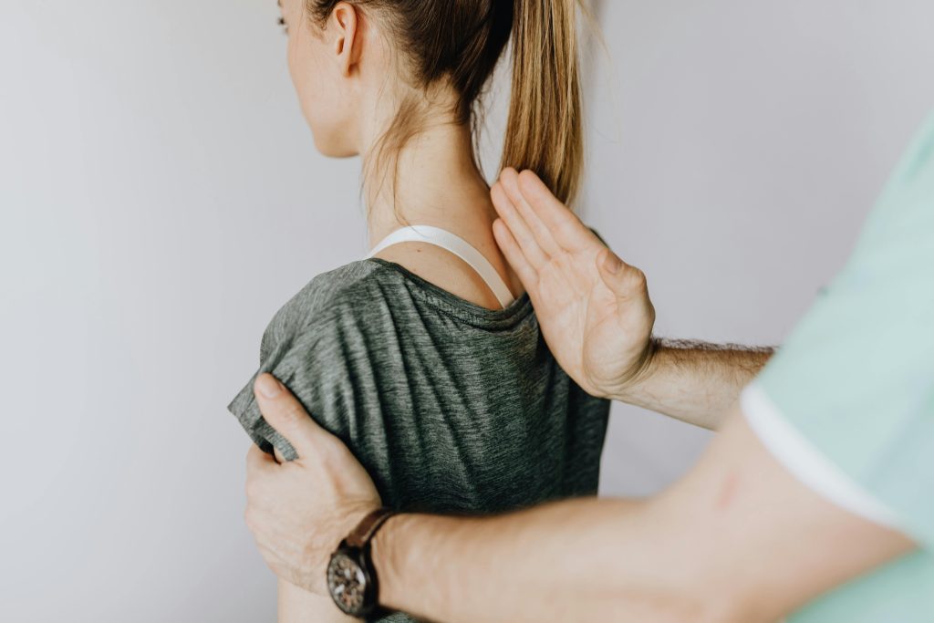 Home Back view of crop unrecognizable osteopath in uniform and wristwatch checking up back of slim female patient in casual wear on white background